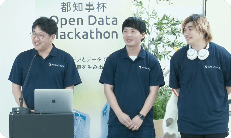 写真：都知事ハッカソンの様子。インフォ・ラウンジの男性社員が3名揃いの紺色のユニフォームを着用している